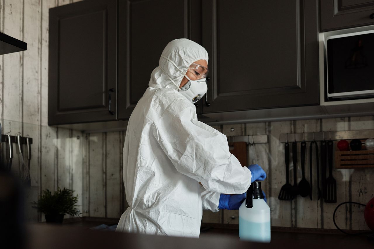 services-06 Healthcare worker in PPE uses disinfectant spray in a kitchen setting during COVID-19 pandemic.